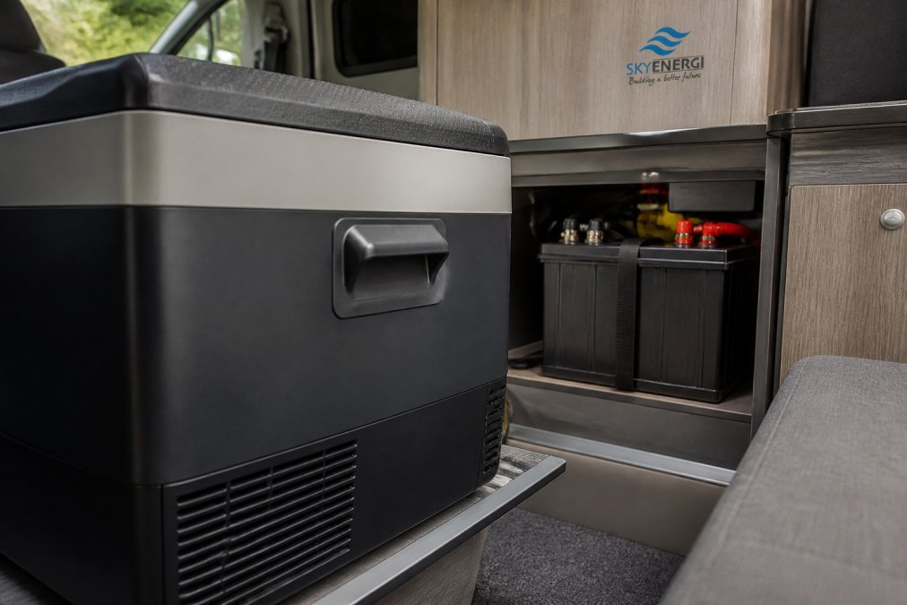 Close-up of a 12V compressor fridge installed in a campervan interior, showing the unit operating with battery terminals visible in the background