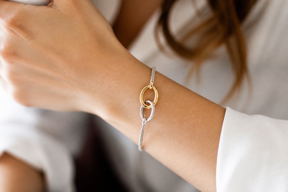 Close-up of a woman's wrist wearing a delicate symbolic mixed metal bracelet with interlocking silver and gold bands, soft natural light highlighting the metallic details against her skin