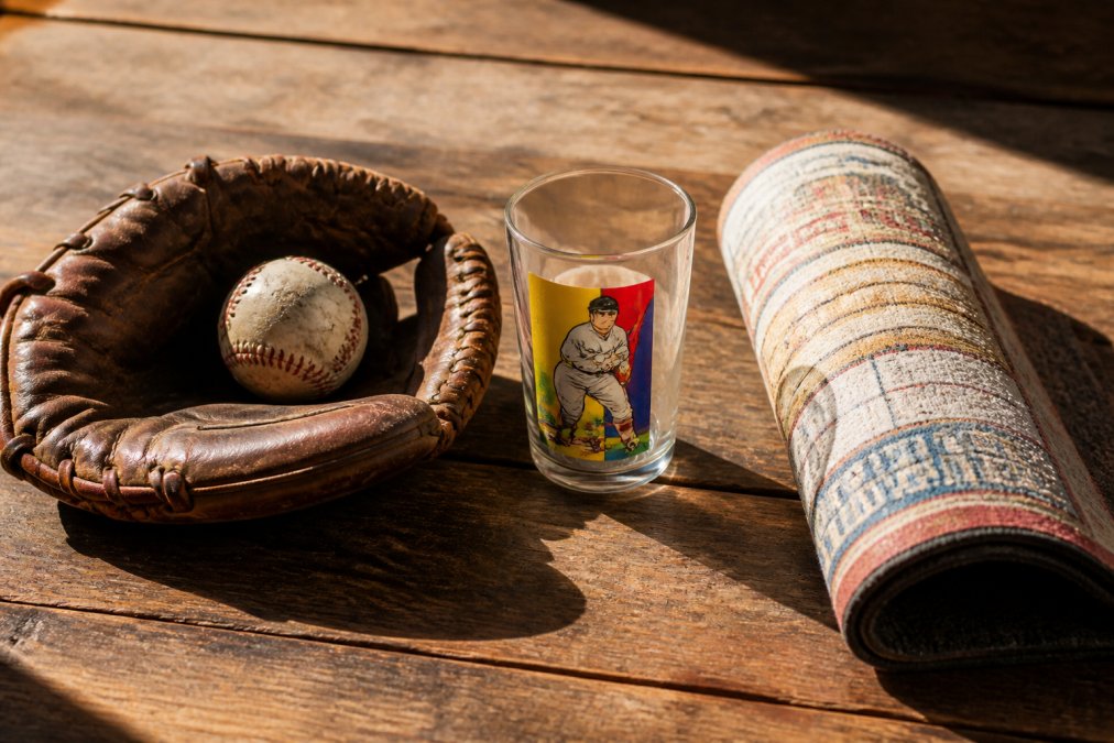 A flat lay of vintage sports memorabilia gifts on a rustic wooden table, including a baseball mitt, a retro ticket doormat rolled at the edge, and a clear 16oz mixing glass with a colorful 1930s baseball card design, lit by warm afternoon sunlight