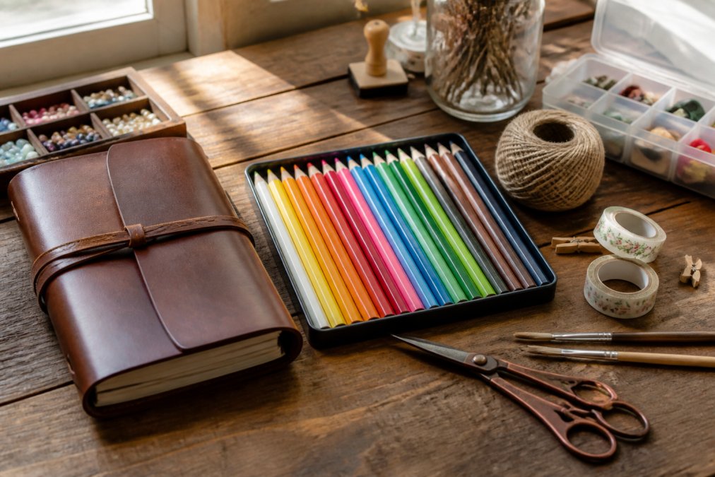 A flat lay photo of various hobby supplies including a vegan leather journal, colored pencils, and craft tools arranged on a wooden table with soft natural window light