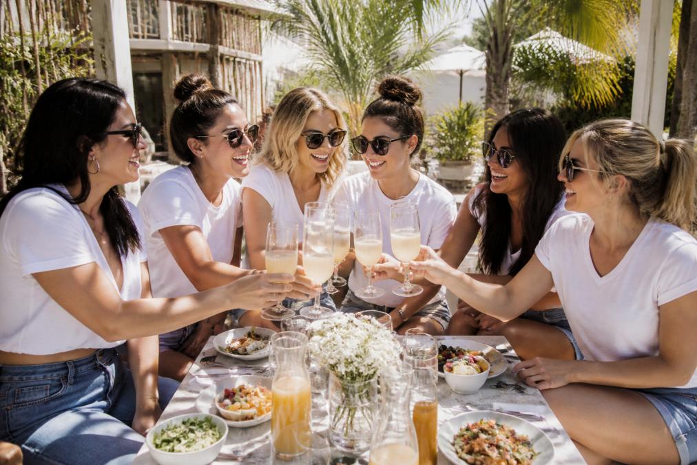 A joyful group of women wearing matching bachelorette shirts, laughing together at a sunny outdoor brunch, holding drinks and posing for a photo