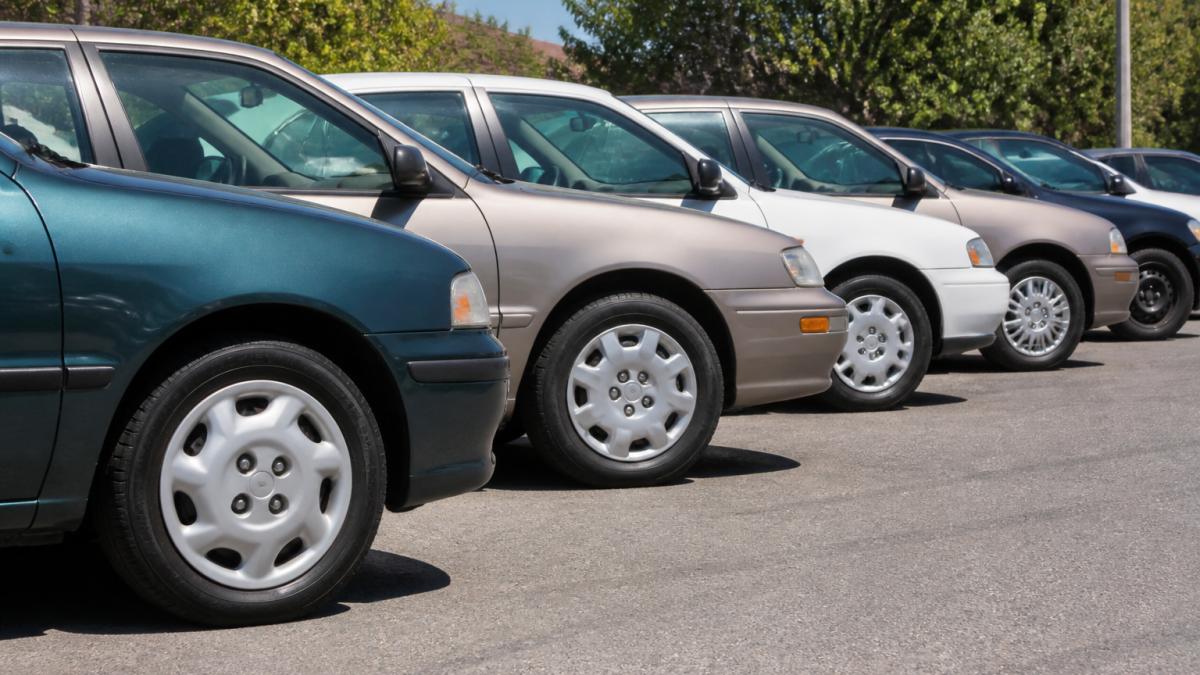 A row of parked older sedans and compact cars in a sunny parking lot, each showing a different 15-inch wheel cover style, with one steel wheel visible without its cover