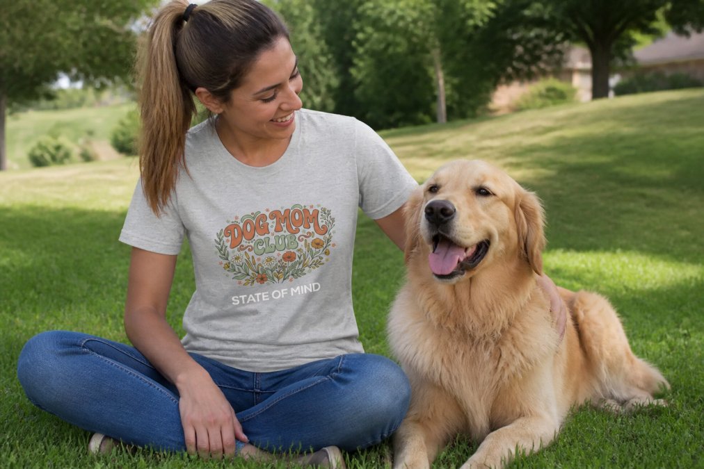 A dog mom wearing a personalized dog graphic tee while sitting on grass with her golden retriever, both looking happy and relaxed in natural sunlight