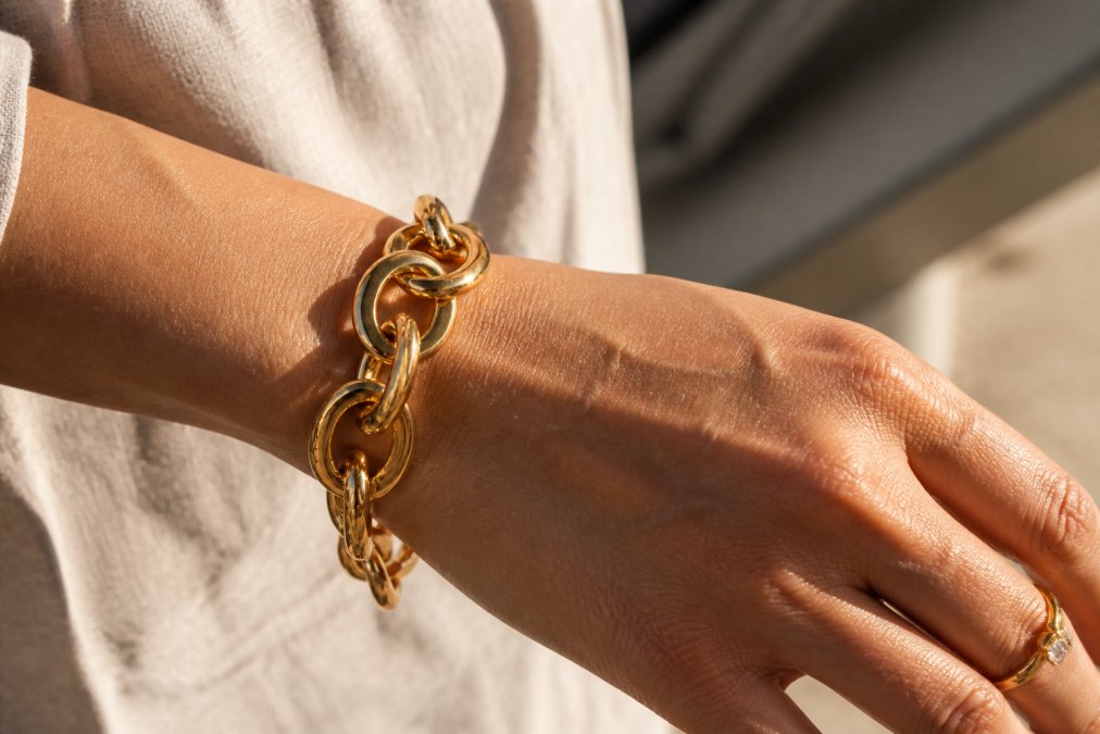 Close-up of a woman's hand wearing an elegant 18k gold statement bracelet, showing the rich saturated color and fine craftsmanship of the metal against her skin, with natural sunlight highlighting the metal's depth