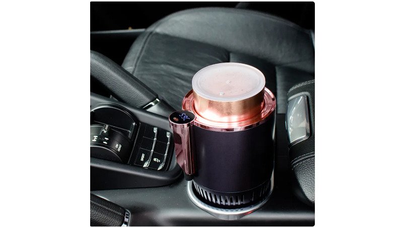 A close-up of a driver's hand placing a stainless steel coffee mug into the smart car cup warmer/cooler mounted in the center console cup holder, the blue digital temperature display glowing clearly against the dark interior
