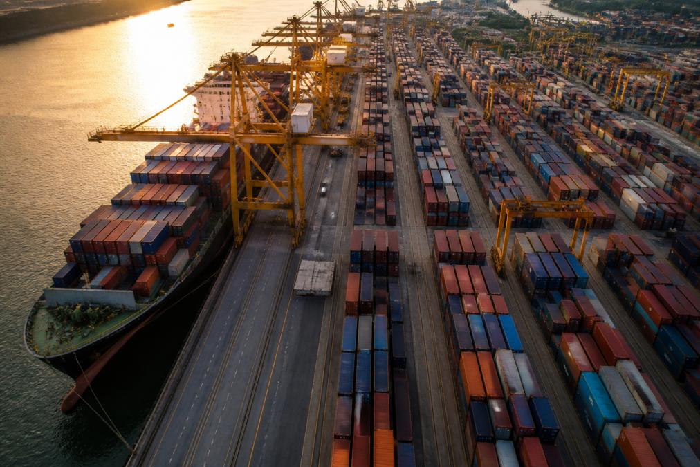 Aerial view of a busy container port at golden hour, rows of stacked 20 ft shipping containers in red and blue stretching toward a massive cargo ship being loaded by gantry cranes