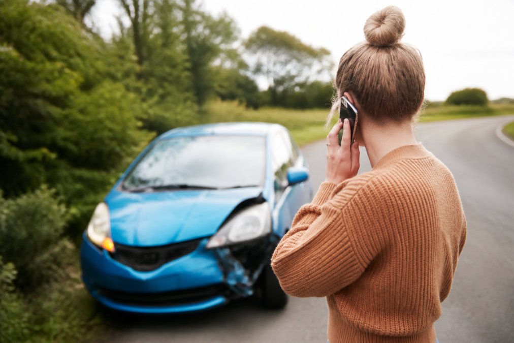 Driver standing safely outside a damaged vehicle on the roadside during daylight, using a mobile phone to call for help, with hazard lights flashing on the vehicle in the background