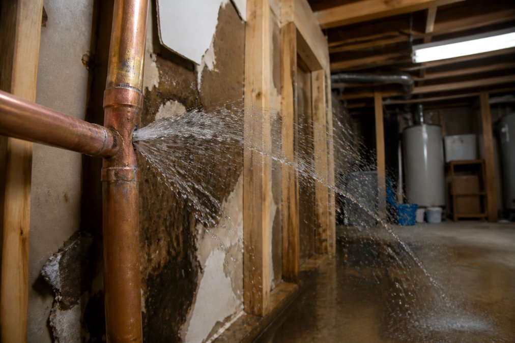 Close-up of water spraying forcefully from a burst copper pipe, with visible water damage to drywall and wooden framing in a residential basement, fluorescent work lights illuminating the scene