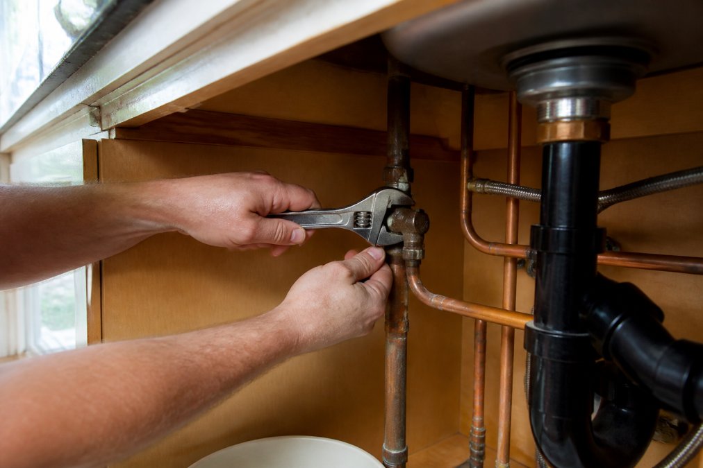 Homeowner's hands gripping a metal shut-off valve wrench beneath a kitchen sink, turning the valve clockwise, with clear access to the valve and surrounding copper piping visible in natural daylight