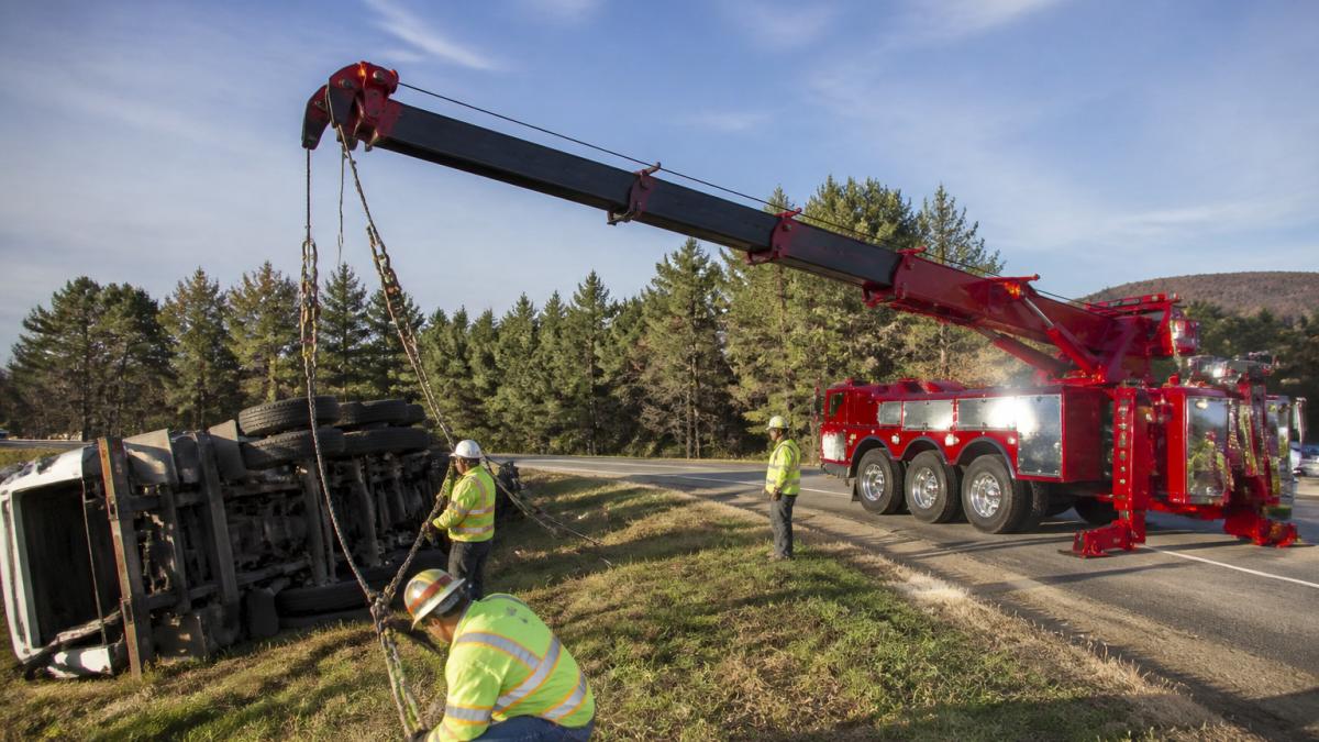 Professional heavy-duty flatbed tow truck with extended rotating recovery crane and winching rigging positioned at an accident scene with multiple operators securing recovery straps, daytime natural lighting