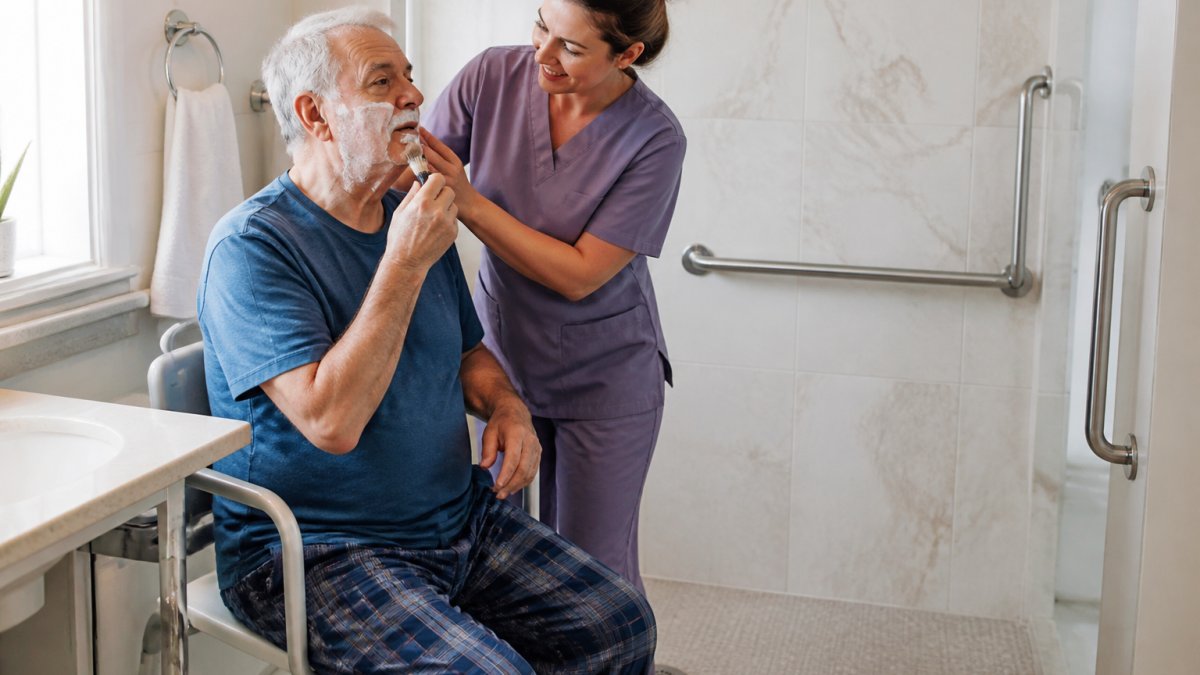 Professional caregiver assisting older adult with grooming in bright, accessible bathroom with grab bars, safety railings, and non-slip flooring while patient maintains active participation
