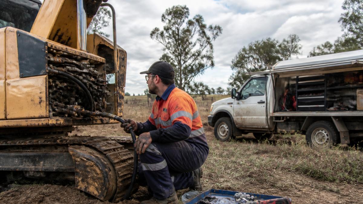 A mobile diesel mechanic in work gear kneeling beside a large earthmoving excavator on a rural Queensland site, hydraulic hose in hand, service truck with open toolbox parked nearby in late afternoon light