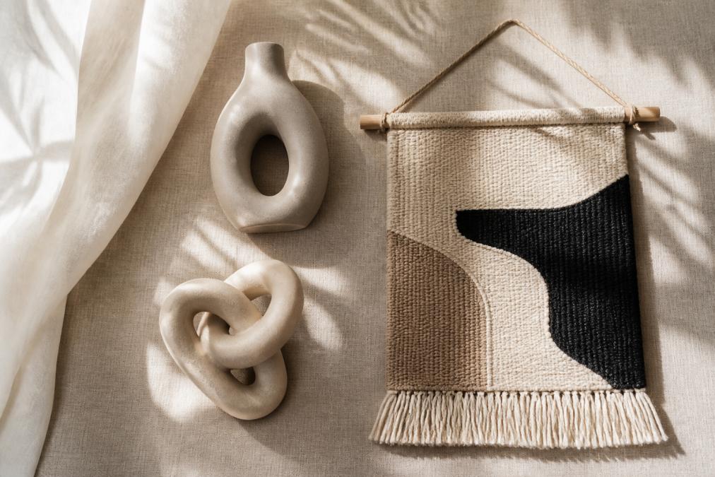 A flat lay of modern decor items including a minimalist ceramic vase, a small abstract sculpture, and a woven wall tapestry arranged on a neutral linen background with soft natural window light