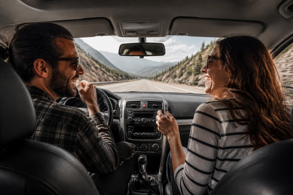Two adults in the front seats of a car smiling and laughing while playing a word game, with a scenic highway visible through the windshield