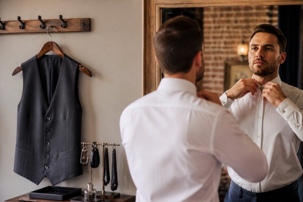 Close-up of a man's hands adjusting the collar of a crisp white dress shirt in front of a mirror, with a tailored waistcoat hanging nearby