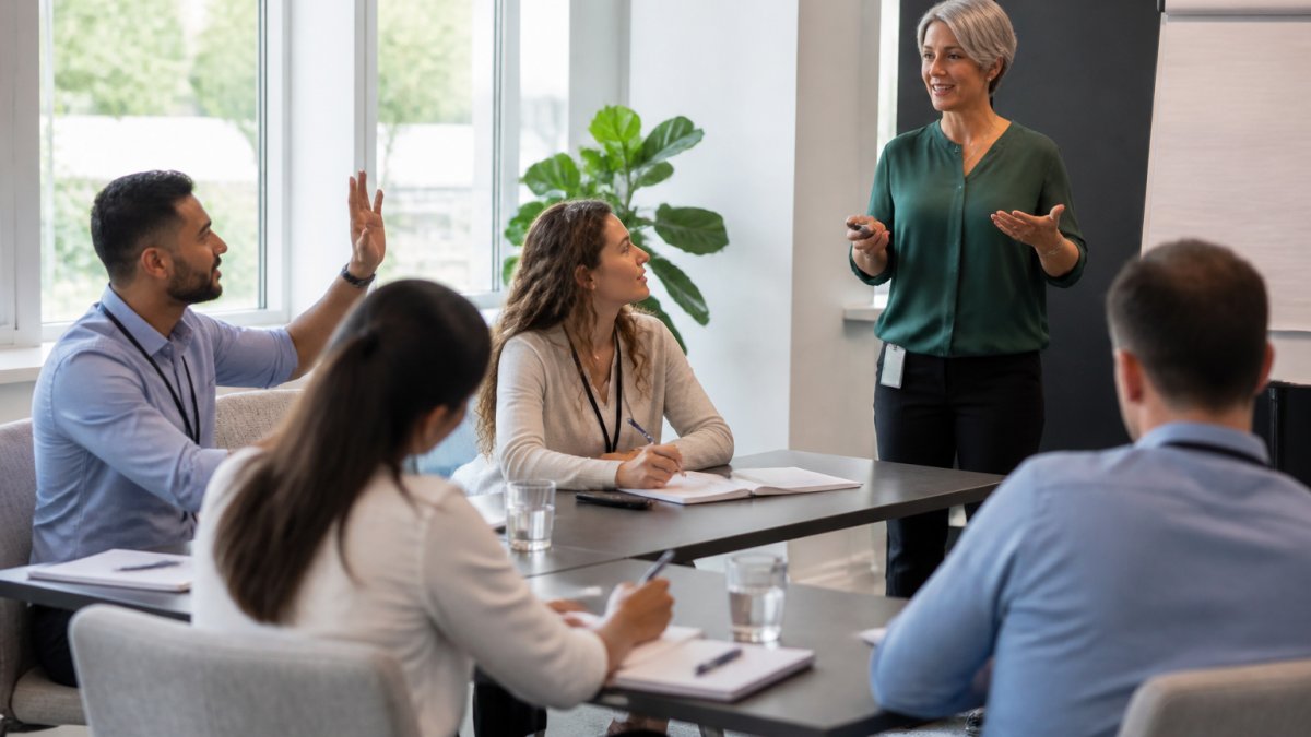 Professional corporate trainer delivering session to engaged participants in modern training room with natural lighting, participants taking notes and asking questions
