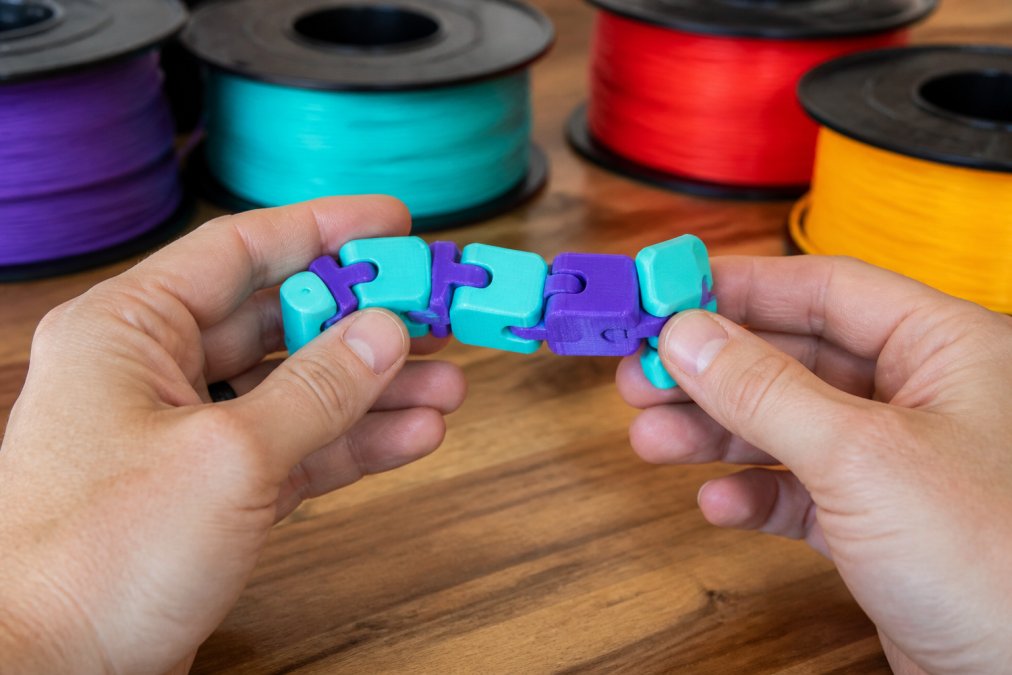 Hands holding a smooth, brightly colored 3D printed fidget toy, showing fine surface detail and articulated moving parts, with spools of filament in different colors softly blurred in the background
