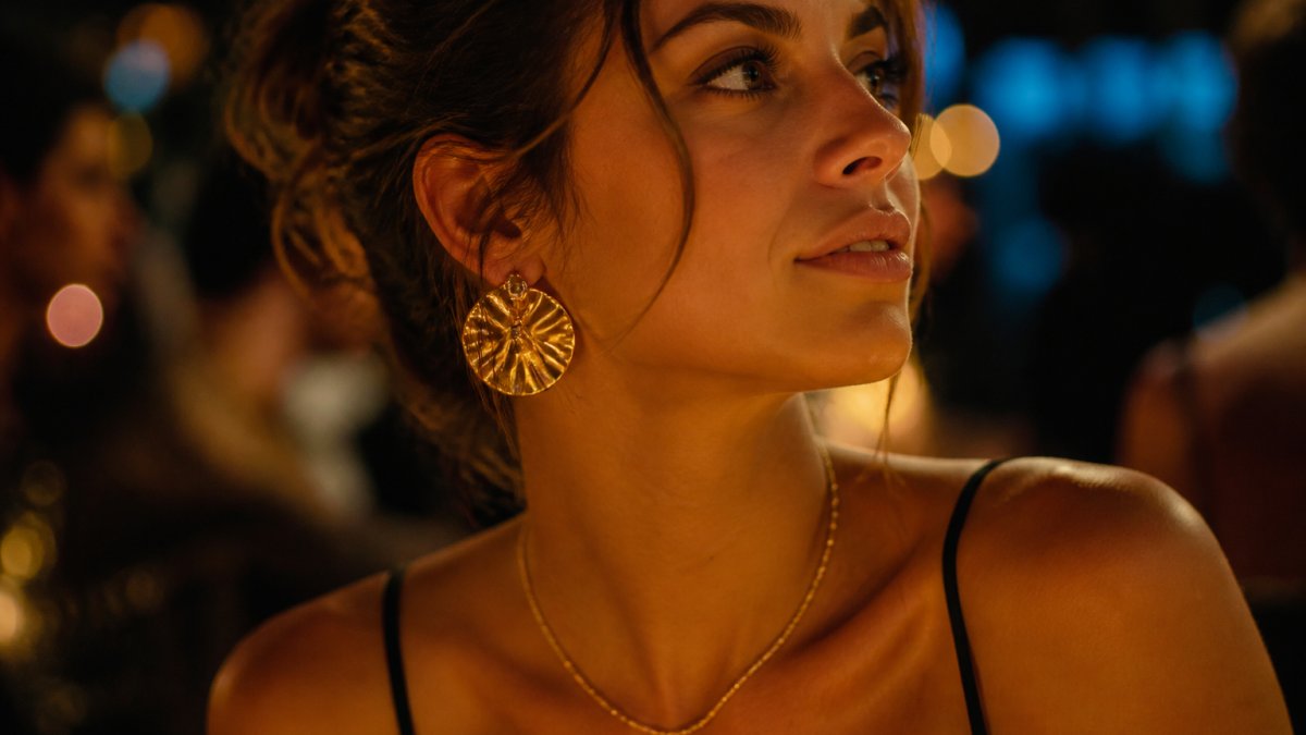 Gros plan d'une femme portant un collier doré fin et des boucles d'oreilles imposantes sous une lumière chaude.
