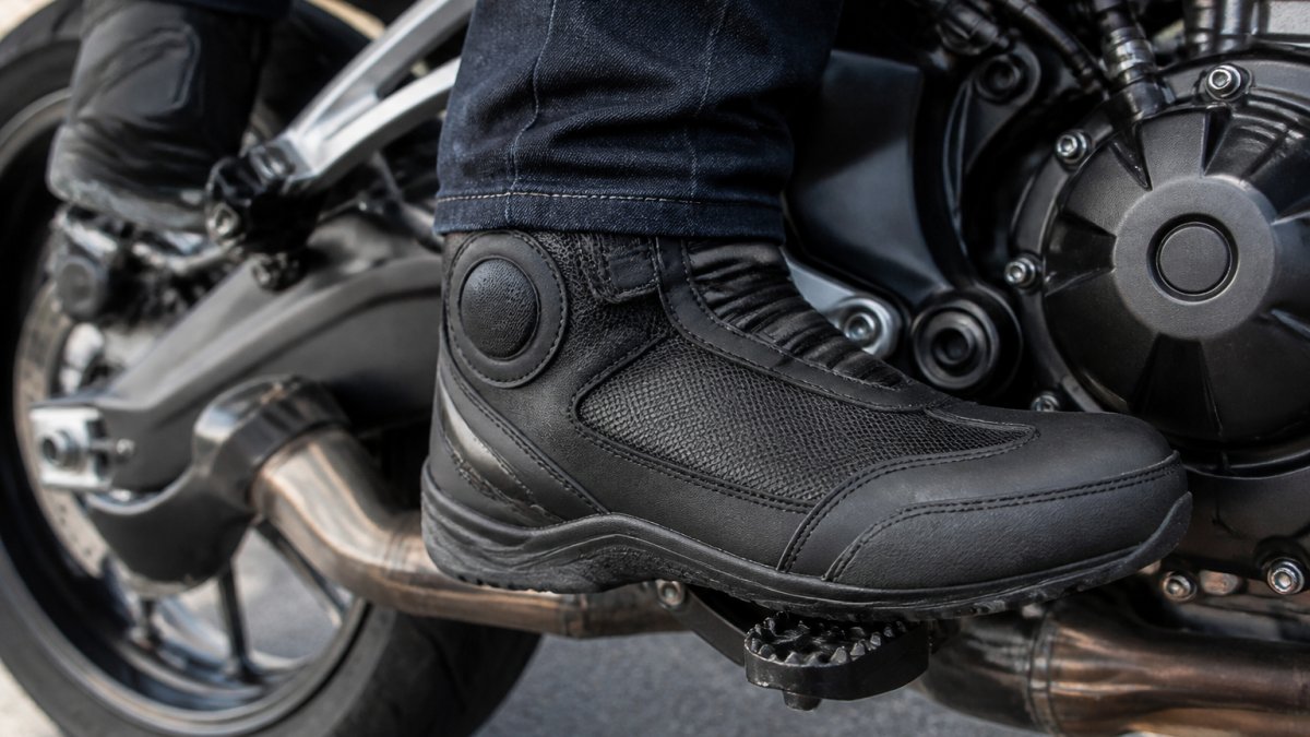 Close-up of a rider's feet positioned on motorcycle foot pegs wearing Alpinestars street boots, showing protective ankle disc details and reinforced toe box in natural daylight