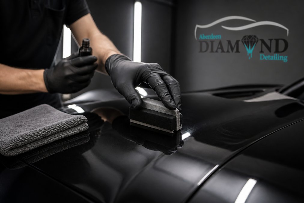 Close-up of a detailer's gloved hand applying a ceramic wax to the glossy black paint of a prestige car, with a microfibre cloth nearby and soft garage lighting