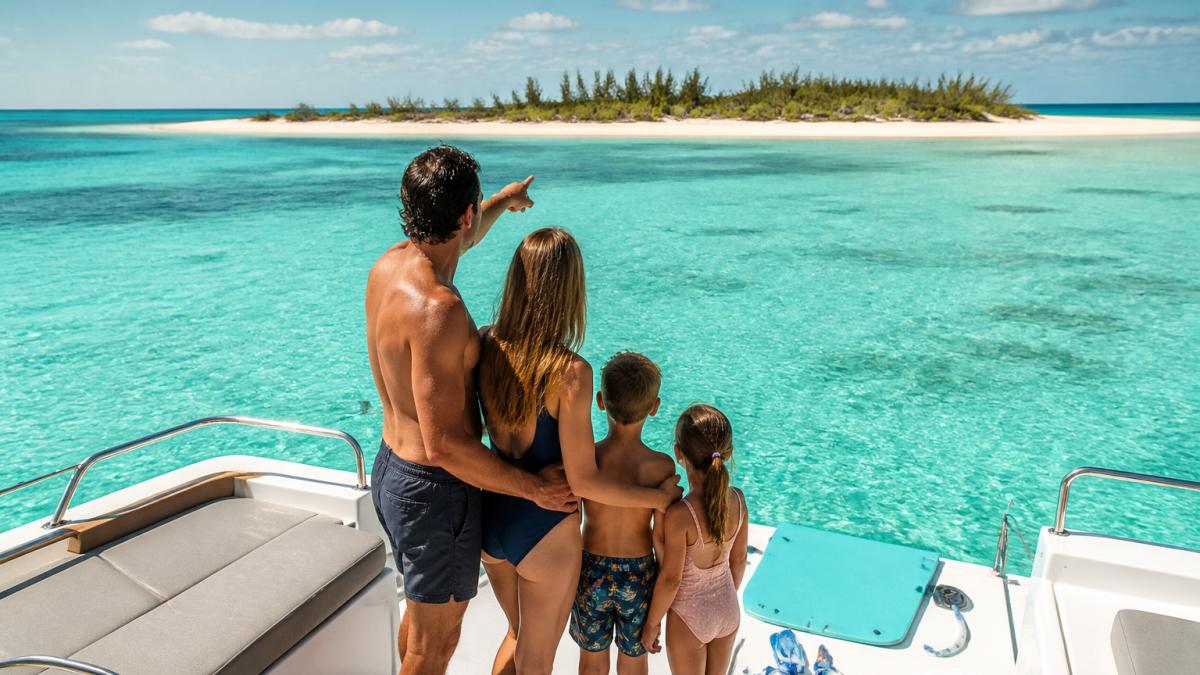 A family with two young children in swimwear on the flybridge of a catamaran anchored in shallow turquoise water, snorkel gear and a floating mat on the aft deck, parents pointing toward a small Bahamian cay in the distance