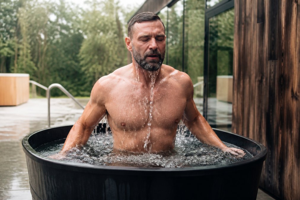 Person emerging from an ice bath with water droplets cascading down shoulders, visible physical response and focused expression in a modern wellness facility