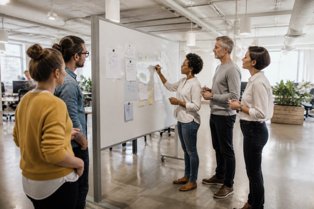 Diverse team of professionals gathered around a mobile magnetic whiteboard partition in a bright open-plan office, actively writing project ideas and discussing strategy together