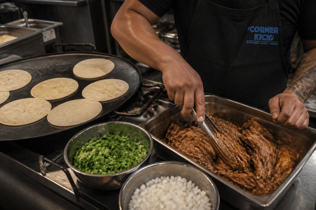 Overhead shot of a taco preparation station with fresh handmade tortillas resting on a griddle, bowls of fresh cilantro and diced white onion nearby, and a chef's hands working with slow-cooked marinated meat in a metal container under warm kitchen lighting