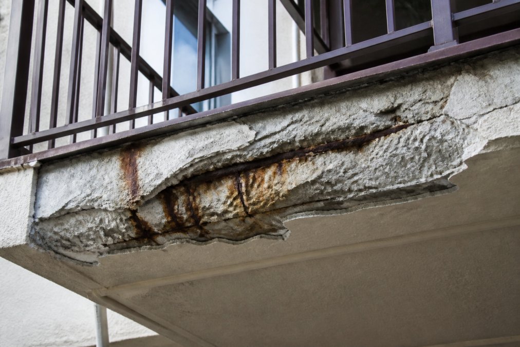 Close-up of a deteriorating balcony edge showing water stains, cracked concrete, and rust streaks on metal reinforcement with visible moisture damage and peeling coating
