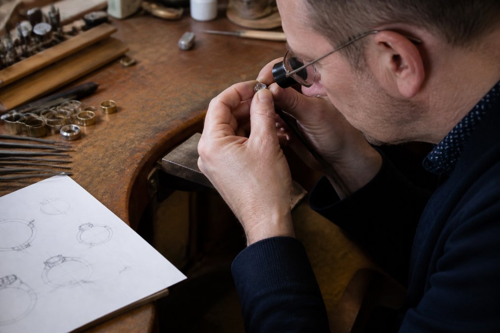 Master jeweller at workbench examining a gemstone under magnifying glass, with jewellery tools, precious metals, and design sketches visible on the work surface in natural studio lighting