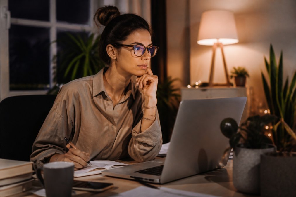Remote worker wearing blue light glasses at desk during evening work, focused on monitor with proper posture and desk setup visible