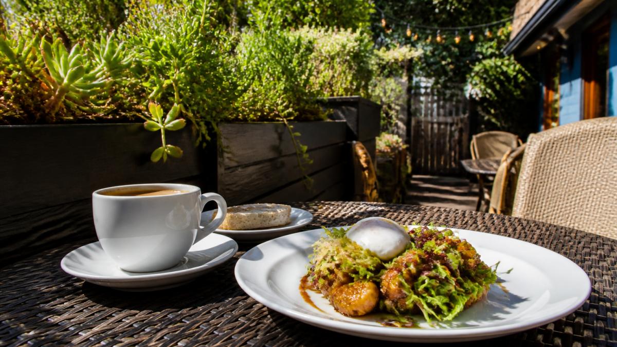A sunlit patio table at a charming Carmel-by-the-Sea restaurant, set with a plate of Asian fusion breakfast dishes and a cup of coffee, with string lights and greenery in the background