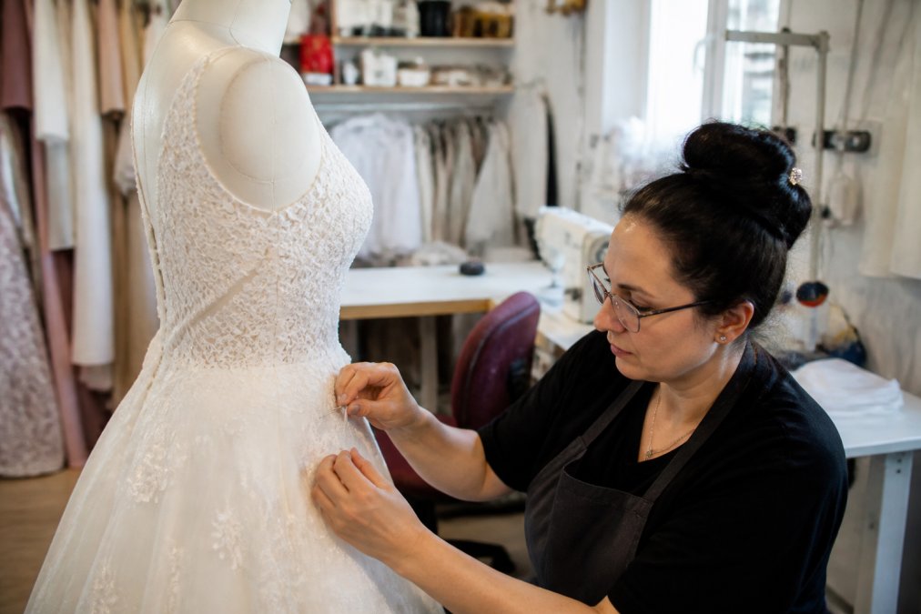 Seamstress pinning the hem of a white lace wedding dress on a mannequin in a bright tailoring studio