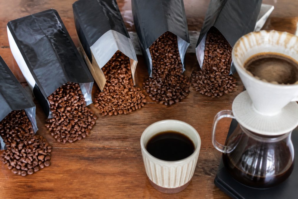 A flat-lay photo of several open coffee sample bags with roasted beans spilling out, alongside a ceramic pour-over brewer and a cup of freshly brewed coffee on a wooden table