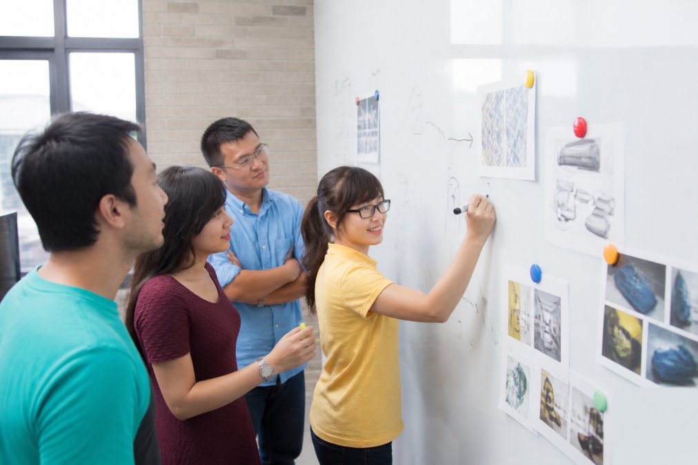Creative team collaborating around a wall covered in white magnetic wallcovering, with hand-drawn sketches in dry-erase marker and colour-coded neodymium magnets holding printed design concepts and mood boards