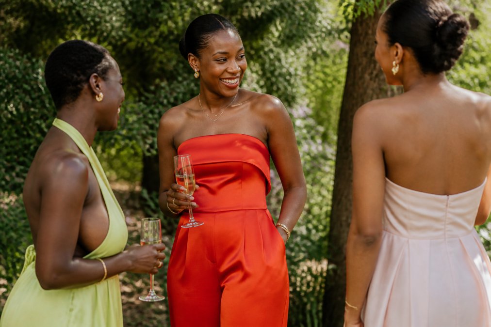 Woman wearing a vibrant persimmon-orange satin jumpsuit with clean strapless neckline, standing in natural garden setting with soft afternoon sunlight illuminating the fabric's luxurious drape and sheen