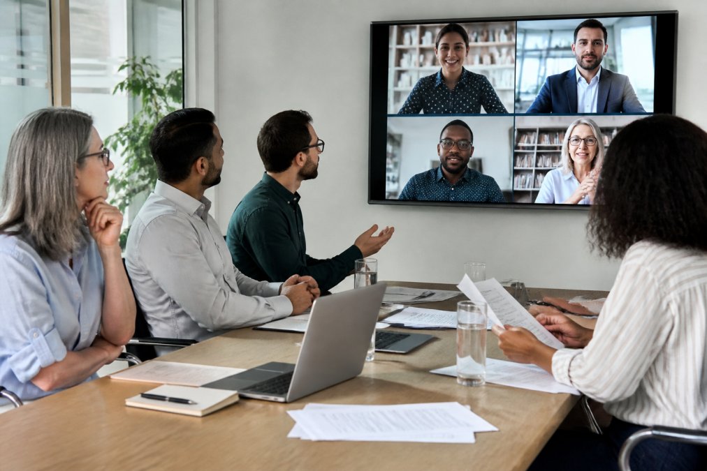 A diverse team of professionals in a hybrid meeting setup, some seated at a conference table in an office with laptops, others visible on a large screen engaged in discussion with attentive body language, natural office lighting