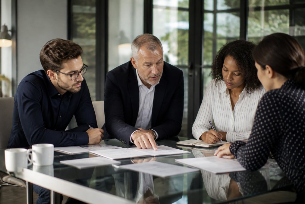 A senior executive leading a focused strategy discussion with three team members around a glass table in a modern office, pointing at a document while others take notes