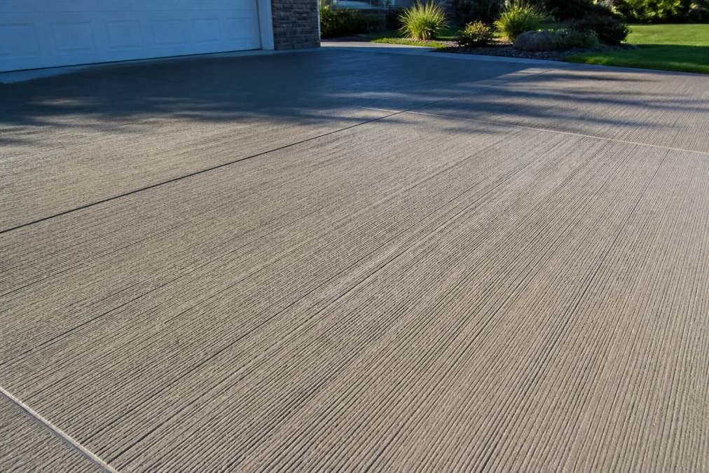 Close-up of a freshly finished driveway with a broom-textured concrete surface showing fine parallel lines of the texture in natural daylight, with morning shadows highlighting the texture depth