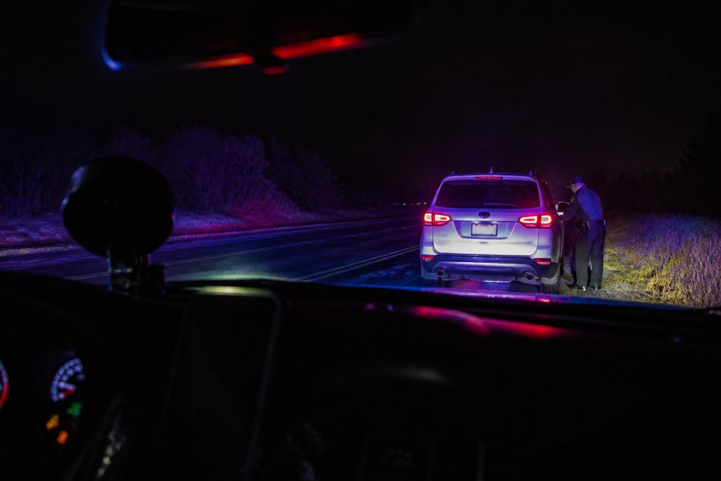 Close-up view from inside a police cruiser at night showing dashboard camera perspective of a vehicle pulled over on a dark road, red and blue emergency lights reflecting off wet pavement