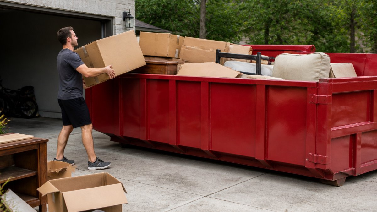 Homeowner loading bulky furniture and cardboard boxes into a roll-off dumpster in a residential driveway, stacking items efficiently with proper technique