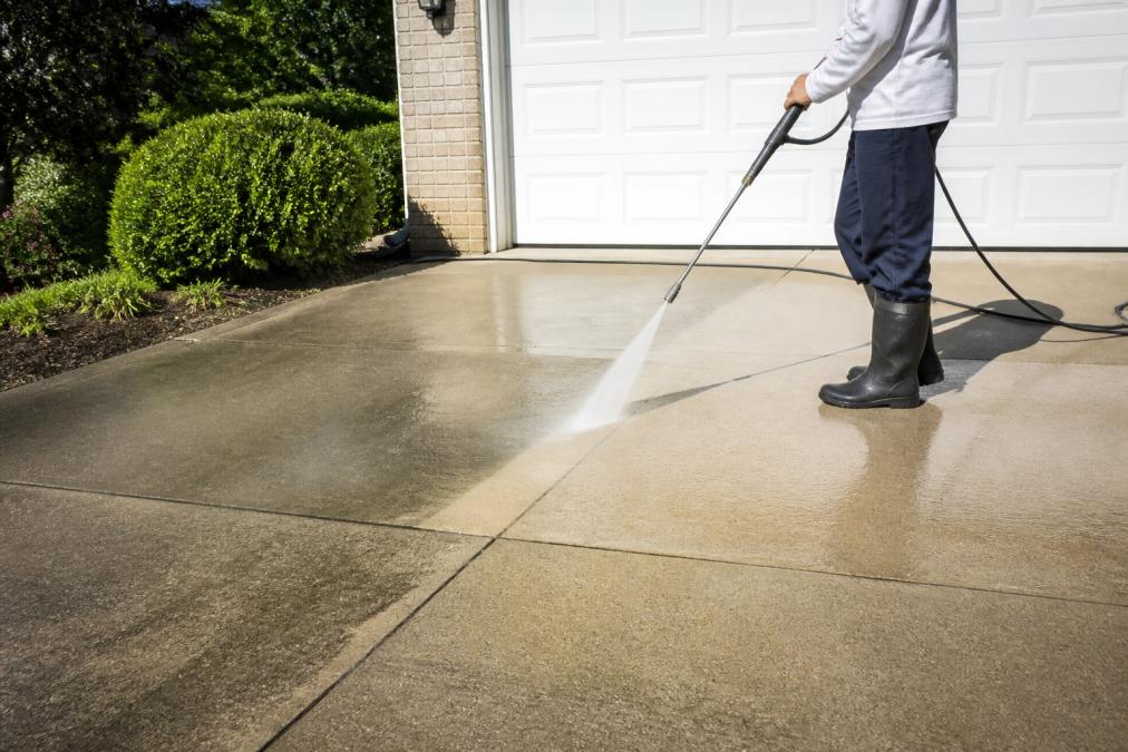 Professional pressure washer cleaning a concrete driveway with high-pressure water spray, showing visible dirt and grime removal in bright daylight