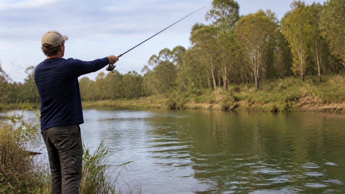 Angler casting a lure along a river bank with trees in background, showing proper casting form and positioning near water edges