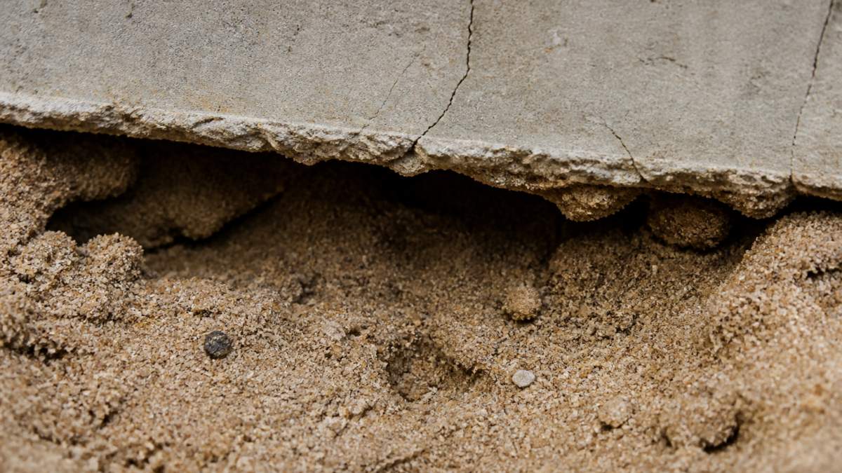 Close-up of sandy soil granules with visible void spaces beneath a concrete foundation slab showing hairline cracks