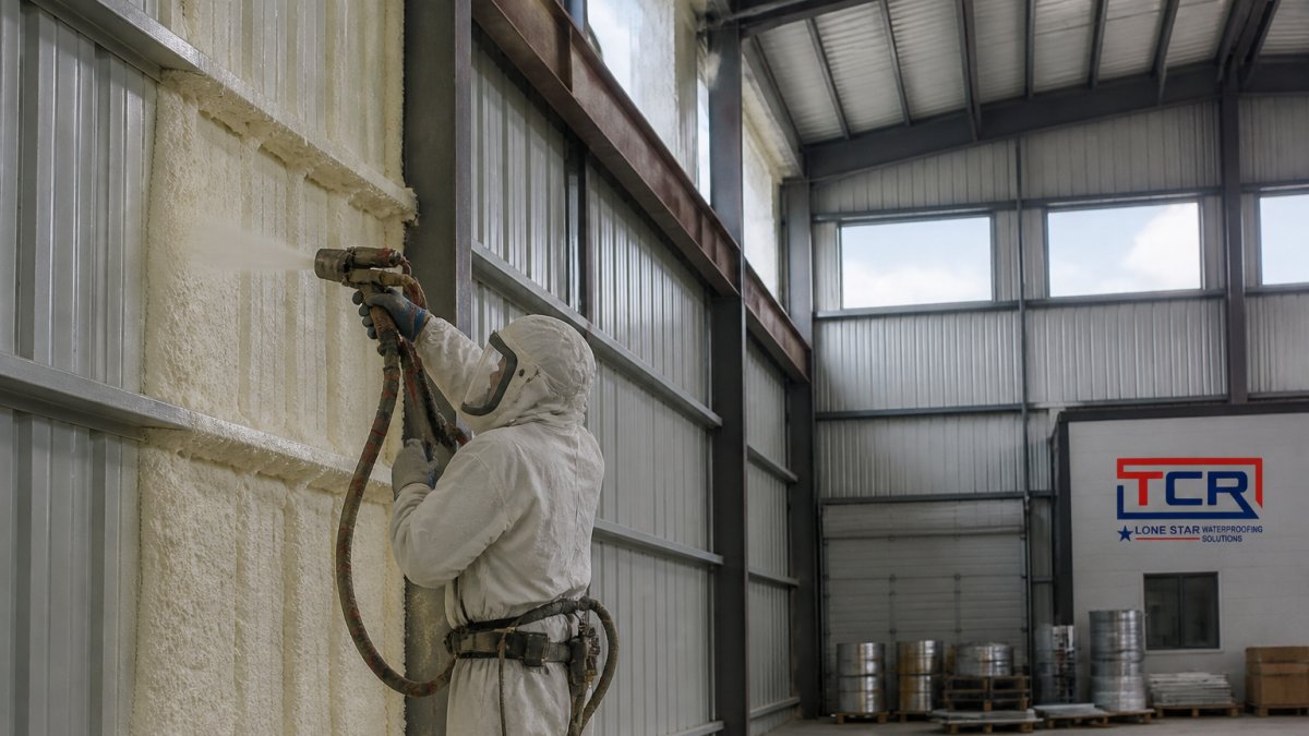 A technician in protective gear spraying closed-cell foam onto the interior metal wall of a large industrial warehouse, with exposed steel purlins and daylight coming through high windows