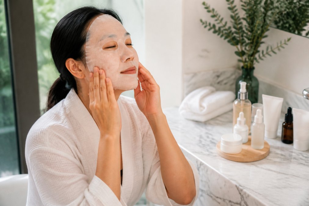 Woman in her mid-30s applying a hydrating face mask at home in soft natural light, with skincare products arranged on a marble bathroom counter, showing a calm self-care moment