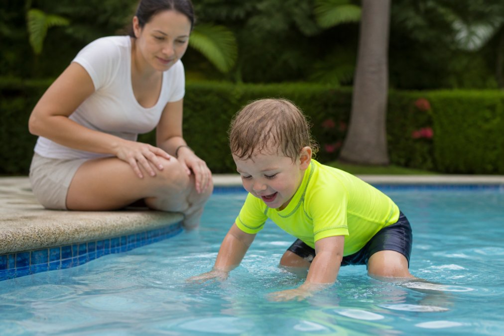 Active toddler in bright neon high-visibility swim set playing in shallow pool water with parent supervising closely from the pool edge