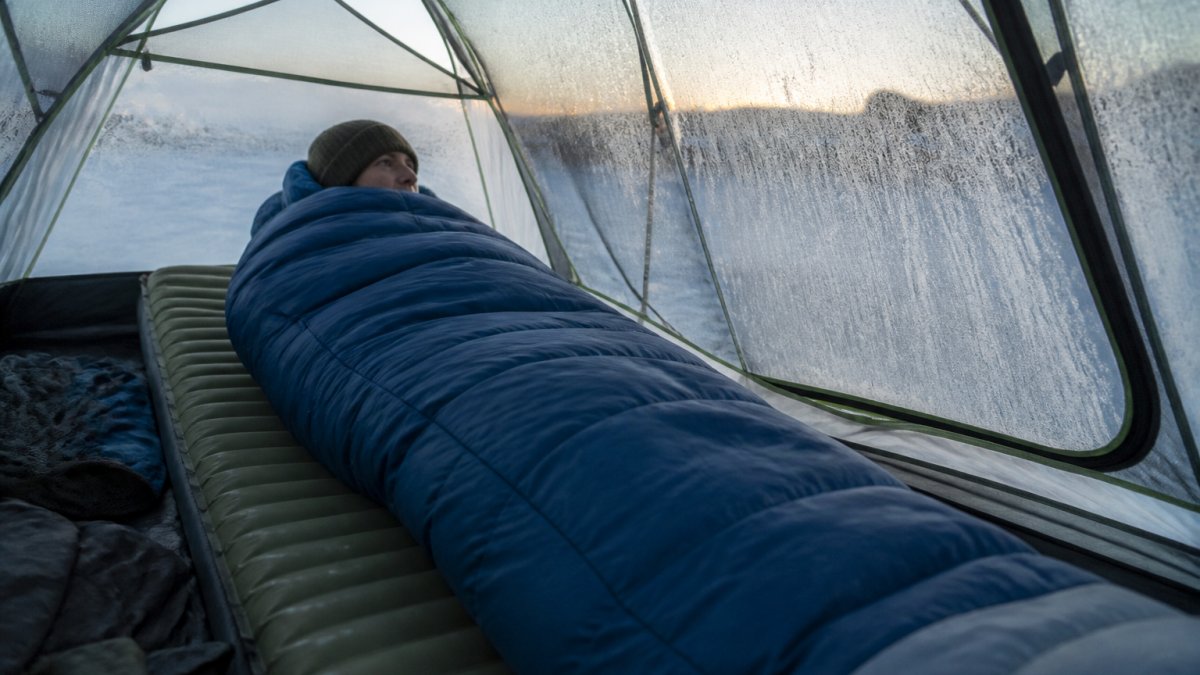A camper in a mummy sleeping bag on an insulated sleeping pad inside a tent, wearing a warm hat, with a draft collar visible and frost on the tent walls at dawn
