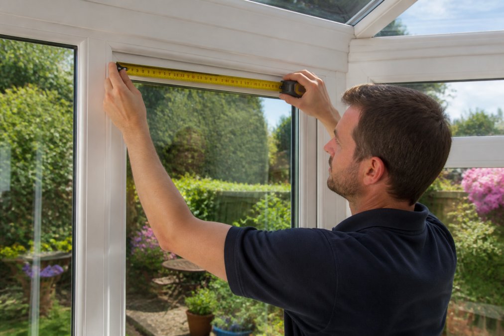 Homeowner measuring a conservatory window with a steel tape measure, showing proper technique at the window recess and uPVC frame edges with natural daylight streaming in