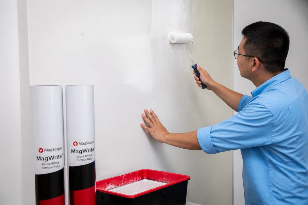 Professional decorator applying magnetic wallcovering to a classroom wall using paste-the-wall method, showing proper surface preparation and adhesive application with tools and materials visible